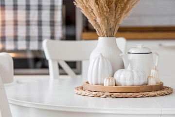 Still-life. Dried pampas grass in a vase, white ceramic pumpkins, a teapot and pumpkin-shaped candles on a white table in the interior of a Scandinavian-style home kitchen. Cozy autumn concept.