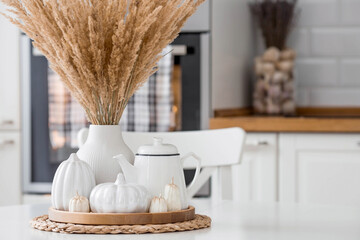 Fototapeta na wymiar Still-life. Dried pampas grass in a vase, white ceramic pumpkins, a teapot and pumpkin-shaped candles on a white table in the interior of a Scandinavian-style home kitchen. Cozy autumn concept.