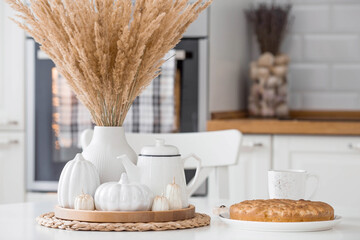 Obraz na płótnie Canvas Still-life. Dried pampas grass in a vase, white ceramic pumpkins, a teapot and pumpkin-shaped candles on a white table in the interior of a Scandinavian-style home kitchen. Cozy autumn concept.