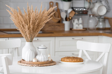Still-life. Dried pampas grass in a vase, white ceramic pumpkins, candles and apple pie charlotte on a white table in the interior of a Scandinavian-style home kitchen. Cozy autumn concept.