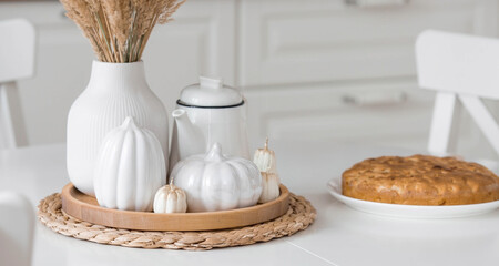 Still-life. Dried pampas grass in a vase, white ceramic pumpkins, candles and apple pie charlotte on a white table in the interior of a Scandinavian-style home kitchen. Cozy autumn concept.
