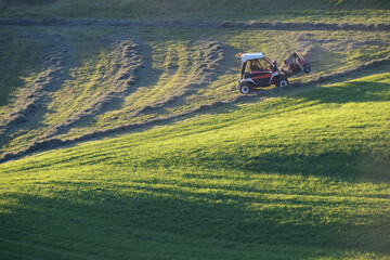 Mountain farmer rakes on the green steep slope in the evening light, slope tractor in harvest use
