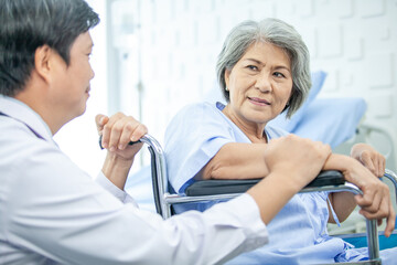Obraz premium Asian doctor supporting and cheering up senior patient in wheelchair talking, smiling in comfort at hospital. Healthcare and medicine concept.