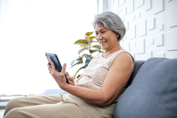 Asian senior woman using smartphone for video call with happy mood smiling and laughing on sofa at home.