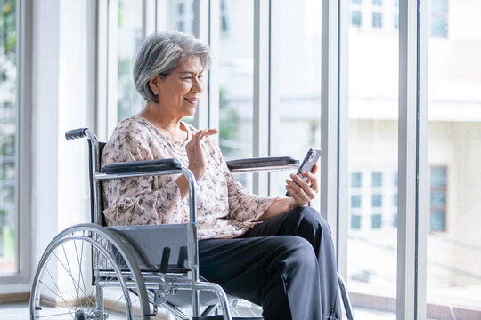 Happy elderly asian woman sitting on wheelchair and using smartphone for video call at home.