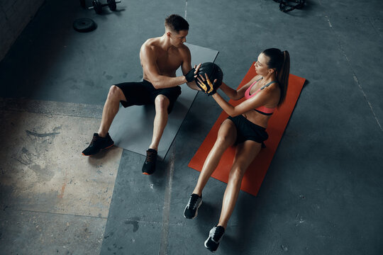 Top view of young fit couple exercising with medicine ball while sitting on exercise mats in gym