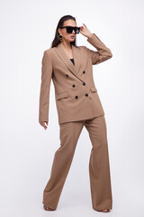 Fashion photo of a beautiful elegant young woman in a pretty oversize brown and beige suit, jacket, pants, stylish sunglasses posing over white background. The hair is gathered back, dark brunette.