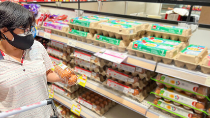 Asian woman wearing a mask while considering buying eggs in supermarket whose prices on the rise...