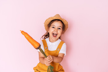 Little girl in orange overalls holding carrot in her hands against pink background.