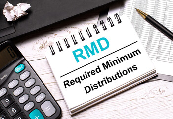 On a light-colored office table is a computer, a black calculator, a pen, and a notebook with the text RMD Required Minimum Distributions