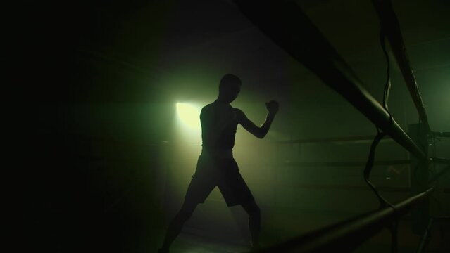 Lightweight fighter training inside the boxing ring. Silhouette of a boxer or kickboxer athlete punching the air, shadow fight in a empty foggy gym