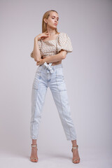 High fashion photo of a beautiful elegant young woman in pretty blue denim jeans, beige top with floral pattern posing on white, soft gray background. Slim Figure, Blonde. 