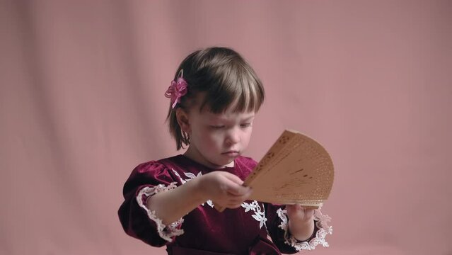 Cute little girl in an vintage burgundy dress is playing with a wooden fan. The concept of children theater productions and developing acting circles. Coral background