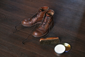 Pair of brown leather stylish boots, footwear cream mink oil and shoe brush on wooden floor, shoe care products