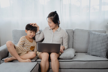 A middle-aged Asian single mom is working on a laptop on the sofa in the living room of the house. The son ran and jumped onto the sofa and sat next to his mother, begging her for attention.