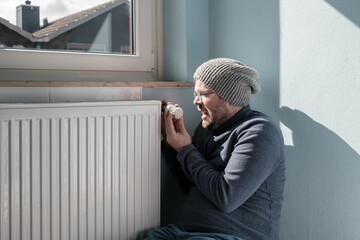 Irritated man in warm clothes turns the radiator thermostat to minimum. Concept of crisis and...