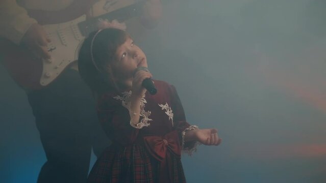 Little girl on stage in vintage dress, she sings into microphone and dances, her father plays an electric guitar. Color music is shining and smoke is billowing. Family time, creativity and hobbies