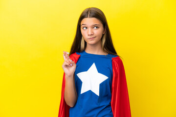 Little caucasian superhero girl isolated on yellow background with fingers crossing and wishing the best