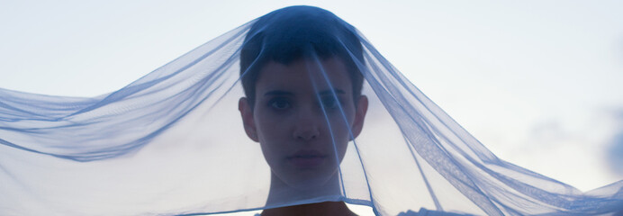 Young woman in white dress hiding her face behind white transparent veil in the nature