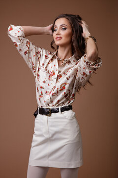 High fashion photo of a beautiful elegant young woman in a pretty patterned shirt, white short skirt, tights, brown shoes, accessories posing over beige background. Studio Shot. Portrait. 