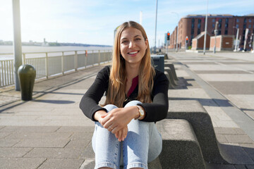 Young beautiful woman sitting relaxed on Liverpool waterfront on sunny day, England - obrazy, fototapety, plakaty