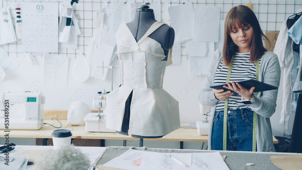 Wall mural Young female fashion designing entrepreneur is working with tablet while checking clothing pinned to tailoring dummy. Modern technologies in clothes manufacturing concept.