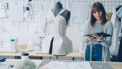 Young female fashion designing entrepreneur is working with tablet while checking clothing pinned...