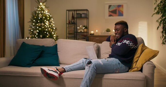 Cheerful guy of African appearance sits on couch and watches comedy movie on laptop. He laughs out loud. The guy is wearing warm sweater, ripped jeans and sneakers. Behind shimmering Christmas tree.