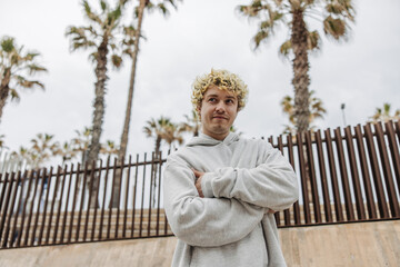 Handsome young caucasian male crossed his arms on chest with palm trees in background. Calm blonde man dressed grey sweater. Concept weekend.