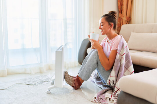 Happy Smiling Satisfied Joyful Young Woman With Plaid And Knitted Socks Drinking Hot Tea And Warming Near Electric Heater At Home