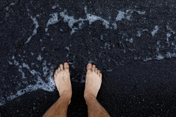 Legs on the background of the beach. Top view of bare feet and legs in fine black pebbles with the movement of waves coming to the feet - foaming sea texture. The concept of summer holidays and vacati