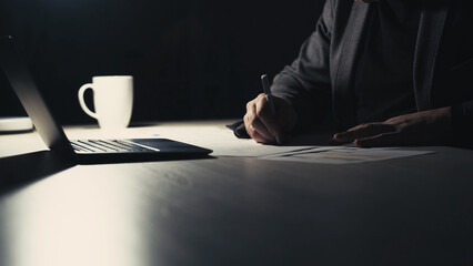 Overwork night. Writing man. Distance work. Unrecognizable business male making notes on paper sitting desk in dark room interior.