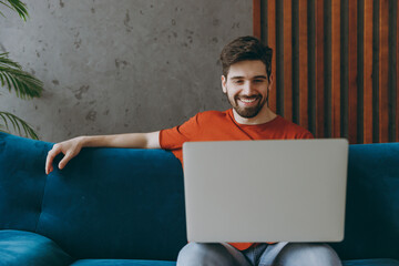 Young IT man in red t-shirt hold use work on laptop pc computer sit on blue sofa couch stay at home...