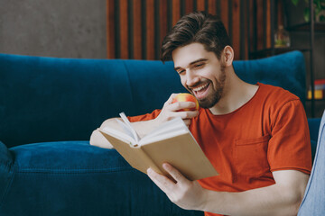 Young calm man 20s wears red t-shirt bite apple read book novel sit on blue sofa couch stay at home hotel flat rest relax spend free spare time in living room indoors grey wall. People lounge concept.