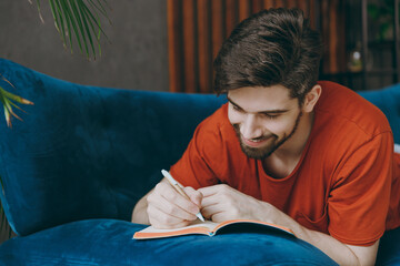 Young calm man wear red t-shirt writing memories in notebook diary lay down on blue sofa stay at...