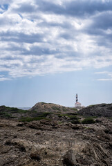 Fototapeta na wymiar distant image of the Favaritx lighthouse, with a rock formation around it on a cloudy summer day, vertical, copy space, Menorca, Balearic Islands, spain