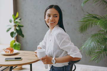 Side view young fun calm tranquil latin woman 30s wear white shirt hold cup drink coffee look aside sit alone at table in coffee shop cafe restaurant indoors. Freelance mobile office business concept.