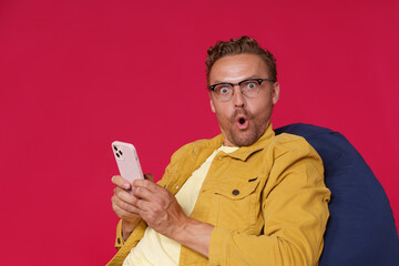 Excited, shocked handsome man in eye glasses hold smartphone in hands looking at camera with WOW look wearing yellow denim jacket sitting on bag chair isolated on red background