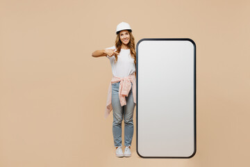 Full body young employee laborer handyman woman in white t-shirt helmet point on big blank screen area mobile cell phone isolated on plain beige background Instruments accessories for renovation room. - Powered by Adobe