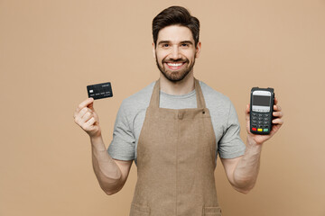 Young man barista barman employee wear brown apron work in coffee shop hold bank payment terminal...