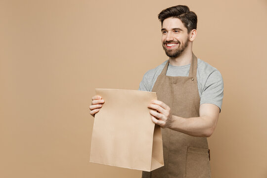 Young Man Barista Barman Employee Wear Brown Apron Work In Coffee Shop Give Craft Paper Bag For Takeaway Food Products Isolated On Plain Pastel Light Beige Background. Small Business Startup Concept.