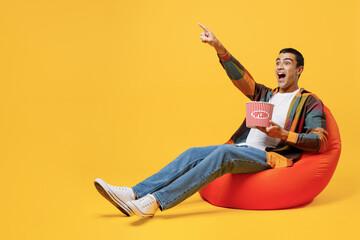 Full body shocked young middle eastern man he wear casual shirt white t-shirt sit in bag chair watch movie hold takeaway popcorn bucket point finger aside isolated on plain yellow background studio.