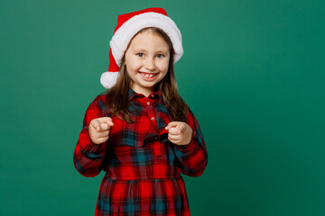 Merry little child kid girl 6-7 years old wear red dress Christmas hat posing point index finger camera on you motivating encourage isolated on plain dark green background Happy New Year 2023 concept