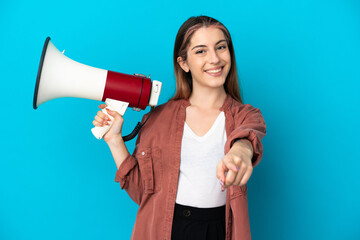 Young caucasian woman isolated on blue background holding a megaphone and smiling while pointing to...