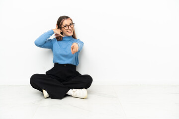 Obraz premium Young Caucasian woman sitting on the floor making phone gesture and pointing front
