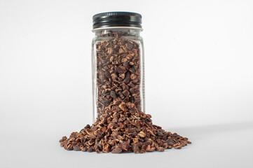 Full of organic raw cacao nibs in glass jar with black lid with a little heap in front of it. Isolated on white background.