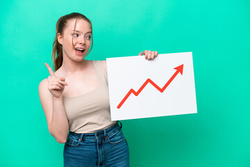 Young caucasian woman isolated on green background holding a sign with a growing statistics arrow symbol and thinking