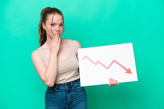 Young caucasian woman isolated on green background holding a sign with a decreasing statistics arrow symbol with surprised expression