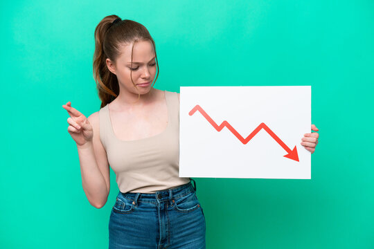 Young caucasian woman isolated on green background holding a sign with a decreasing statistics arrow symbol with fingers crossing