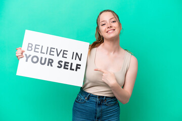 Young caucasian woman isolated on green background holding a placard with text Believe In Your Self and pointing it